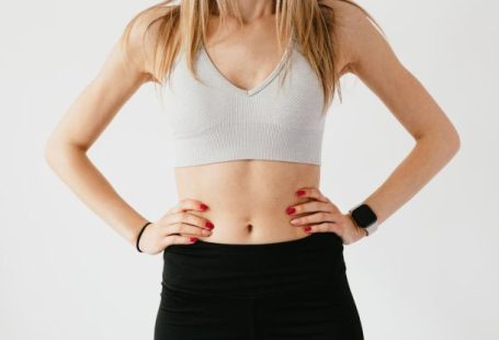 Wearable Fitness Gear - Faceless slim anonymous blond female in sports bra and black leggings in wearable bracelet showing perfect belly on white background while standing with hands on waist