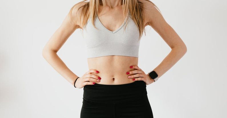 Wearable Fitness Gear - Faceless slim anonymous blond female in sports bra and black leggings in wearable bracelet showing perfect belly on white background while standing with hands on waist